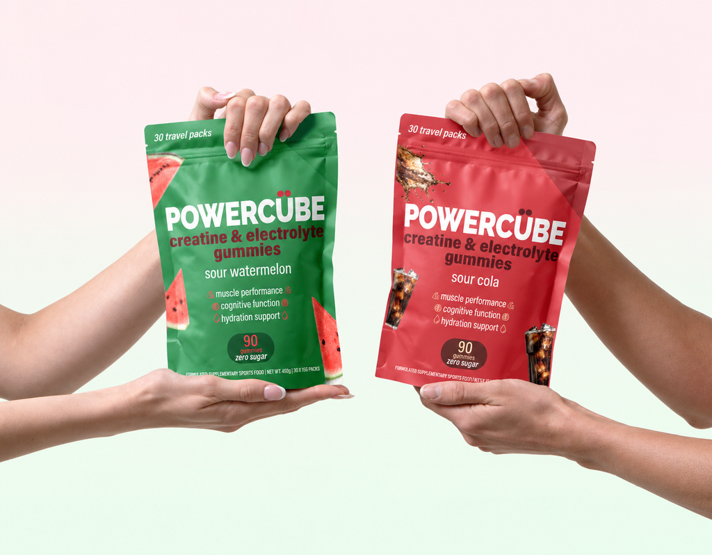 Two hands holding two POWERCUBE creatine and electrolyte gummies pouches, one green sour watermelon flavor and one red sour cola flavor, against a soft pink and light green gradient background.