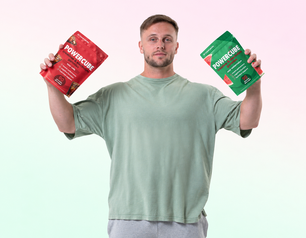 A man holding two pouches of POWERCUBE protein powder, one red and one green, against a soft pastel gradient background.