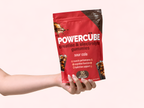 Hand holding a PowerCUBE Sour Cola creatine and electrolyte gummies pouch against a pale pink background.