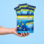 A hand holding a blue retail display box of PowerCube Caffeine Gummies against a light blue background. The display box contains six individual packet packages of gummies, labelled 'blue razz lemonade' with 'POWERCUBE CAFFEINE GUMMIES' on the front.