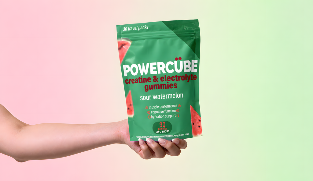 Hand holding a PowerCUBE Sour Watermelon creatine and electrolyte gummies pouch against a light pink and green background.