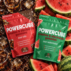 Two stand-up pouches of POWERCUBE creatine and electrolyte gummies are shown on a styled flat-lay with sliced watermelon and dark soda around them. The left pouch is red and labeled sour cola, and the right pouch is green and labeled sour watermelon.